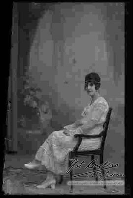 Foto de estudio de una dama que se encuentra sentada en una silla. Luce un vestido largo con bordados, adornos en el cabello y zapatillas con tacón.