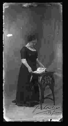 Foto de estudio de una dama que se encuentra parada y observando una revista en una mesita. Lleva puesto una blusa de encaje de color blanco y un vestido largo de color oscuro.