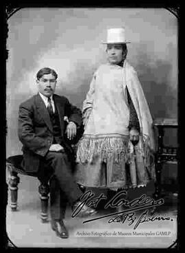 Foto de estudio de una pareja. Un varón de traje, sentado en un sillon junto a una chola que lleva puesto un sombrero blanco de copa alta, aretes, manta bordada, pollera y botines con tacón.