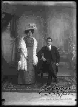 Foto de estudio de una dama que lleva puesto un sombrero, vestido largo y chal de plumas. Se encuentra junto a un varón que viste de traje con chaqueta y se encuentra sentado en un sillón.