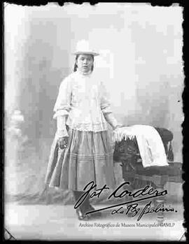 Foto de estudio de una chola con sombrero blanco de copa alta, blusa de manga larga con encajes, pollera y botines con tacón, se encuentra apoyada a un sillón donde esta puesto una manta