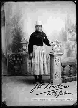 Foto de estudio de una chola que se encuentra parada y apoyada a la columna de una baranda donde esta un jarrón con flores. Lleva puesto un sombrero blanco de copa alta, una chompa tejida de lana con cintas, de color negro, pollera  y botines con tacón