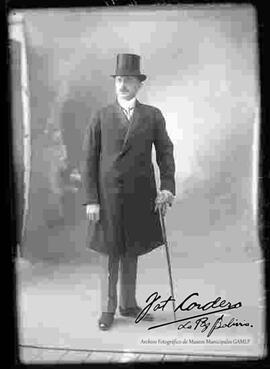 Foto de estudio de un extranjero que lleva puesto: Un sombrero tarro, camisa blanca, corbata, sacón y zapatos guets. Sostiene en mano unos guantes y un bastón.