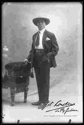 Foto de estudio de un joven que viste de traje y sombrero. Se encuentra parado a lado de un sillón.