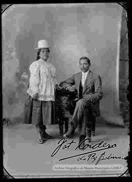 Foto de estudio de una pareja de esposos. Una chola que se encuentra parada, con sombrero blanco de copa alta, blusa ancha, maternal de manga larga, pollera y botines con tacón. Su esposo se encuentra sentado en un sillón vestido de traje y corbata