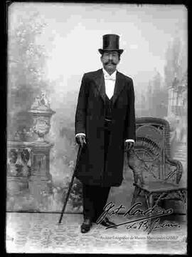 Foto de estudio de un varón de lentes que se encuentra parado a lado de una silla de mimbre. Lleva puesto: Un sombrero tarro, camisa blanca, corbata de gato, chaleco, guantes en las manos y un juego de traje con sacón. Sostiene en mano un bastón.