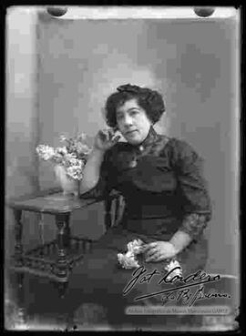 Foto de estudio de una dama que se encuentra sentada y apoyada a una mesita con un jarrón de flores. Presenta un modelo de peinado para cabello corto.