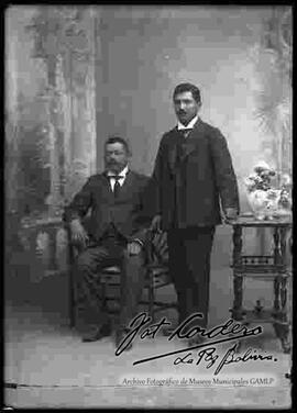 Foto de estudio de dos varones que visten de traje. Uno se encuentra sentado y el otro parado junto a una mesita