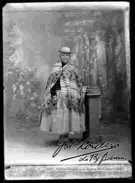 Foto de estudio de una chola anciana que se encuentra parada y apoyada a una pequeña columna. Lleva puesto un sombrero borsalino, manta de alpaca a rayas y con cuadros, pollera con centro sobresalido y botines con tacón.