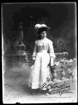 Foto de estudio de una señorita que se encuentra parada junto a un barandal. Lleva puesto un sombrero adornado con flores y pluma; Un vestido de seda largo, sujeto por la cintura con una cinta de seda y  adornado con encajes; Unos guantes en las manos y una cadena en el cuello.