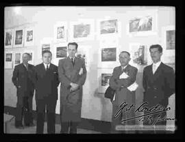 Vista de cinco varones que visten de traje y que se encuentran posando delante de las fotografías de exposición "Foto Cordero Jiménez", en el salón de la cancillería.