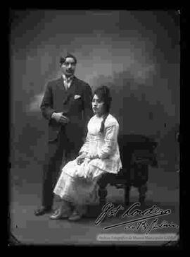 Foto de estudio de una pareja. Un varón que se encuentra parado y que lleva puesto un traje formal y zapatos guets. La dama se encuentra sentada en un sillón y lleva puesto: Un juego de ropa que consta de una blusa, falda larga y una pequeña carterita de color claro.