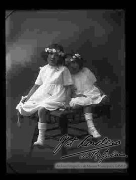 Foto de estudio de dos niñas vestidas de blanco, con flores en el cabello y sentadas encima de una mesita