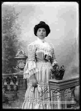 Foto de estudio de una dama que lleva puesto un vestido de fiesta con encajes, sostiene en la mano derecha unos guantes de color blanco y con boton.