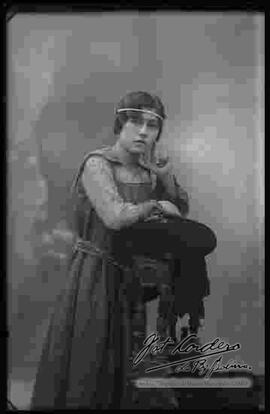 Foto de estudio de una señorita que se encuentra apoyada a un sillón y que presenta un modelo de peinado adornado con una wincha de perlas.