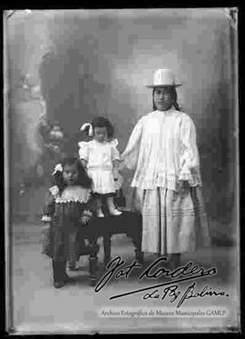 Foto de estudio de una chola que se encuentra parada a lado de sus dos hijas. Lleva puesto un sombrero blanco de copa alta, una blusa suelta de cuello cerrado con manga larga, pollera y botines con tacón