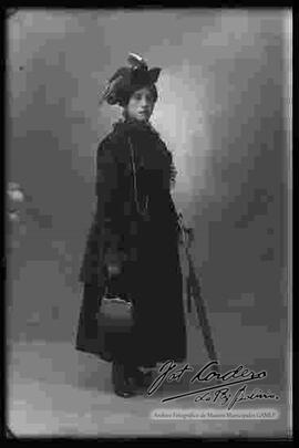 Foto de estudio de una dama que lleva puesto: Un sombrero, un vestido largo de color oscuro y zapatos con tacón, sostiene en las manos una sombrilla y una cartera.