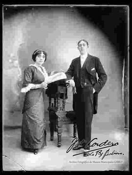 Foto de estudio de una pareja de la clase alta. Una dama que sostiene en mano una revista y que lleva puesto un vestido de fiesta a rayas y un traje con encajes. El varón lleva puesto un traje frac, zapatos guets y sostiene en mano unos guantes.