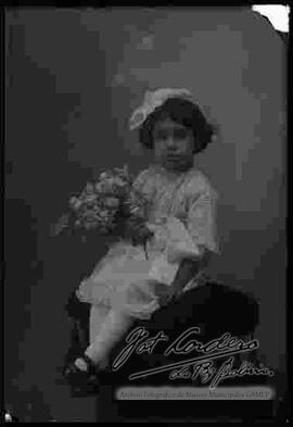 Foto de estudio de una niña que se encuentra sentada en la cabecera o espaldar de un sillón, sosteniendo en mano unas flores