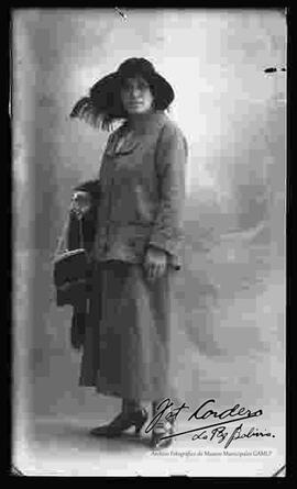 Foto de estudio de una dama que lleva puesto: Un sombrero con pluma, traje con vestido y zapatos de tacón, sosteniendo en mano una cartera y una estola de zorro.