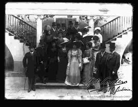 Grupo de damas y caballeros en las escalinatas de una casa. Presentan modelos de sombreros así como también vestimenta.