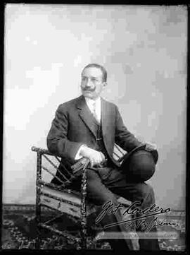 Foto de estudio de un varón de bigote, que se encuentra sentado y viste de traje. Sostiene en mano un sombrero y guantes.