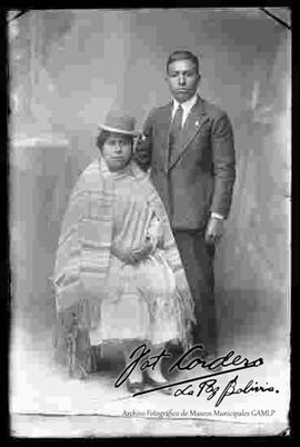 Foto de estudio de una chola junto a su pareja. La mujer se encuentra sentada en una silla, luciendo un sombrero borsalino de color oscuro, manta a rayas o con franjas con sobre una manta, pollera y zapatillas planas.