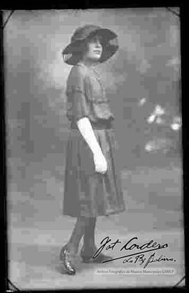 Foto de estudio de una señorita que lleva puesto: Un sombrero, vestido de media manga, guantes blancos en las manos y zapatos de tacón.