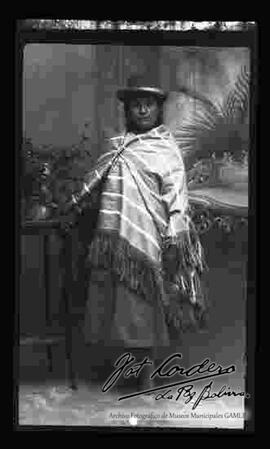Foto de estudio de una chola campesina, la cual se encuentra apoyada a una mesita. Luce un sombrero borsalino de color negro, manta a rayas, pollera y zapatillas planas