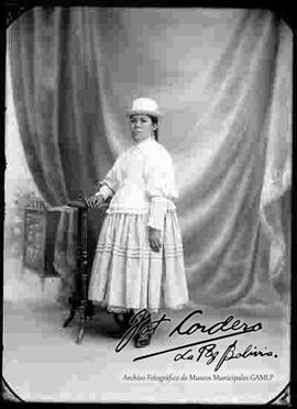 Foto de estudio de una chola que se encuentra parada y apoyada a una mesita. Lleva puesto un sombrero blanco, blusa cerrada de manga larga con encajes de color blanco, pollera con centros sobresalidos y botines con tacón de color negro.