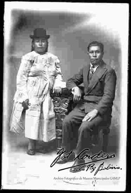 Foto de estudio de una pareja de esposos. Una chola que se encuentra parada con sombrero borsalino color negro, blusa blanca de cuello cerrado, manga larga con encajes, junto a su marido que se halla sentado en un sillón apoyado, con traje y corbata
