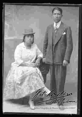 Foto de estudio de una chola que se encuentra sentada junto a su esposo. Luce un sombrero borsalino de color oscuro, blusa de media manga y pollera floreada con botines de tacón