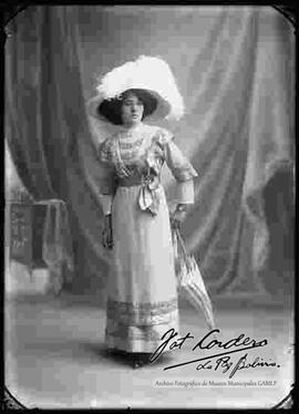 Foto de estudio de una dama. Luce un sombrero, un vestido largo de cuello cerrado, sostenido con una cinta con moño en la cintura y complementado por una sombrilla de sol.