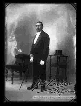 Foto de estudio de un varón que viste de smoking, sosteniendo en la mano derecha guantes blancos y un bastón. Se encuentra parado delante de un sillón y una mesita con tallados donde esta puesto un sombrero