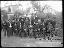 Grupo de personalidades, vestidos de traje y sombrero, posando en el huerto de una casa particular