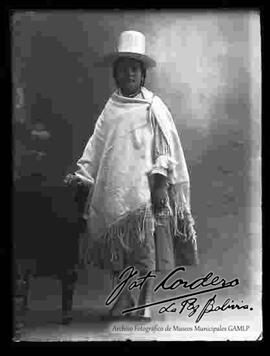 Foto de estudio de una chola que se encuentra parada y apoyada a un sillón. Lleva puesto un sombrero blanco de copa alta, manta,  pollera y botines con tacón