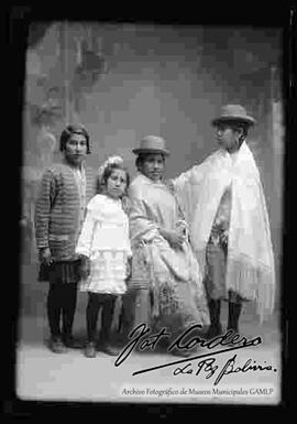 Foto de estudio de dos cholas que se encuentran junto a dos niñas. Ambas llevan puesto sombreros borsalinos, mantas bordadas y polleras. Una con zapatillas y la otra con botines con tacón.