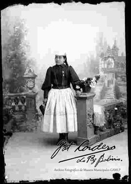 Foto de estudio de una chola que se encuentra parada y apoyada a la columna de una baranda. Lleva puesto un sombrero blanco de copa alta, una cartera en mano, blusa negra cerrada de manga larga, pollera y botines con tacón