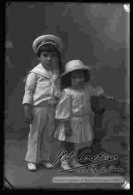 Foto de estudio de una niña y un niño (hermanitos), parados junto a una baranda.