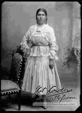 Foto de estudio de una chola sin sombrero que se encuentra parada y apoyada a una silla. Lleva puesto una blusa con encajes de manga larga, pollera y botines con tacón.