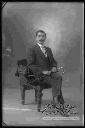 Foto de estudio de un varón que se encuentra sentado en un sillón. Lleva puesto un traje  formal, sosteniendo en manos un bastón.