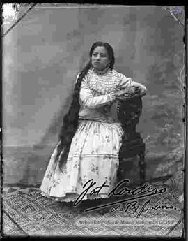 Foto de estudio de una chola que se encuentra sentada en un sillón y con el cabello suelto. Lleva puesto una blusa cerrada, pollera floreada y botines con tacón