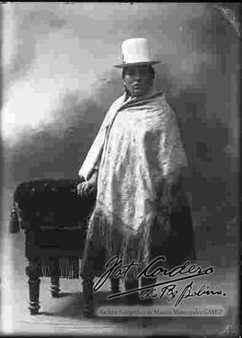 Foto de estudio de una chola que se encuentra parada y apoyada a un sillón. Lleva puesto un sombrero blanco de copa alta, manta bordada, pollera y botines con tacón