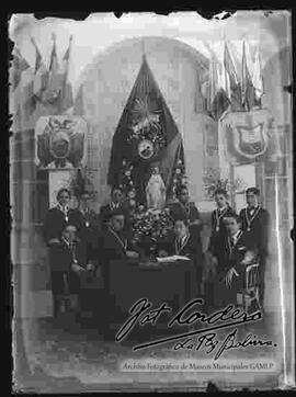 9 estudiantes y 1 cura del colegio San Calixto, con medallas en el cuello, con el fondo  de un estandarte, la imagen de una virgen,  el escudo de Bolivia y escudo de La Paz en el interior de un salón