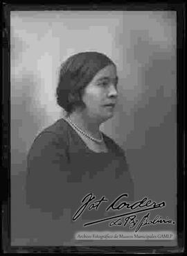 Foto de estudio de medio cuerpo de una dama que se encuentra de perfil. Lleva puesto un collar de perlas y un vestido de color oscuro