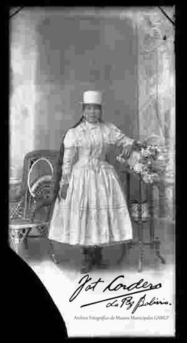 Foto de estudio de una chola que se encuentra parada y apoyada a una mesita con flores. Lleva puesto un sombrero blanco de copa alta, una blusa con encajes de cuello cerrado y manga larga, una pollera y botines con tacón