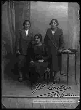 Foto de estudio de tres damas (hermanas). Una de ellas se encuentra sentada en una silla y las otras dos se encuentran paradas lado a lado