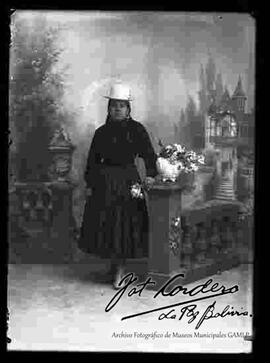 Foto de estudio de una chola que se encuentra parada y apoyada a la columna de una baranda donde esta un jarrón con flores. Lleva puesto un sombrero blanco de copa alta, chompa de lana, pollera de color negro y botines con tacón.