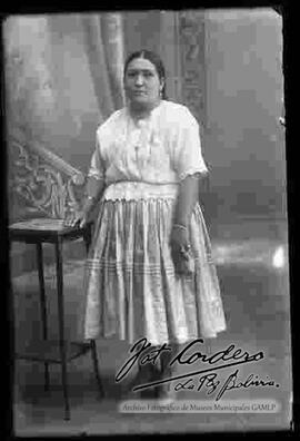 Foto estudio de una chola de cuerpo entero y apoyada a una mesita. Lleva puesto una blusa de manga corta, pollera y botines