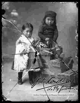Foto de estudio de una niña de pollera y un niño sentado en un bloque de escalones decorativo